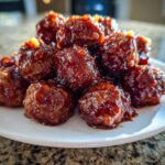 Close-up of a mound of glistening Grape jelly meatballs coated in a thick, dark sauce on a white plate.