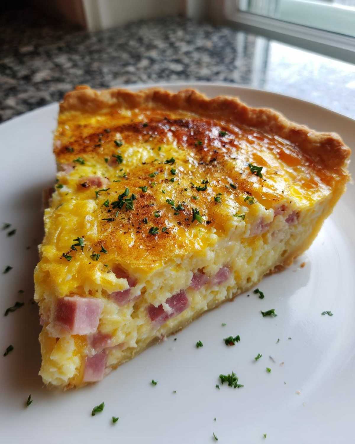 A perfect slice of ham and cheese quiche on a white plate, showing a golden crust and visible chunks of ham.