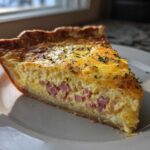 A perfect slice of Ham and Cheese Quiche with a golden crust, filled with egg custard and diced ham, sprinkled with herbs.