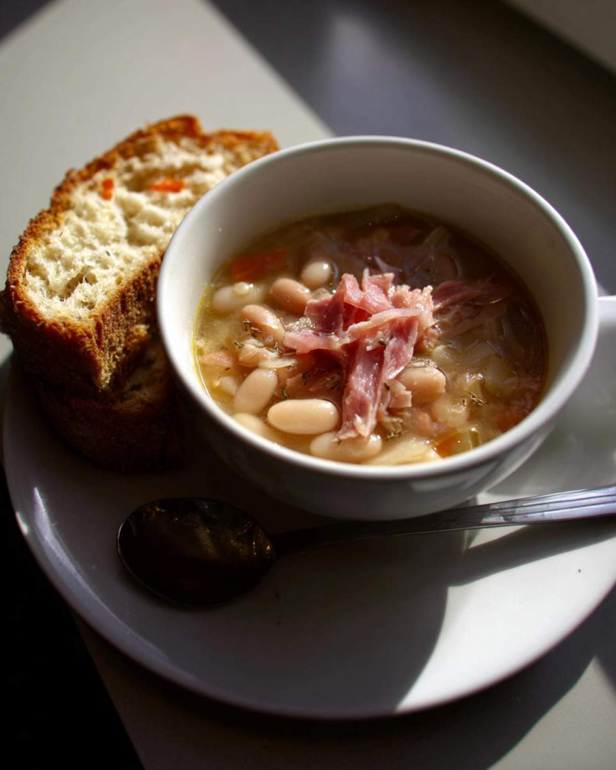 A white bowl filled with hearty bean soup topped with shredded ham, served next to toasted bread.