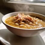 A steaming white bowl filled with thick, hearty bean soup topped with shredded meat, photographed near a window.