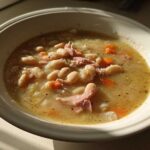 A close-up of a hearty bean soup featuring white beans, shredded ham, and carrots in a savory broth.