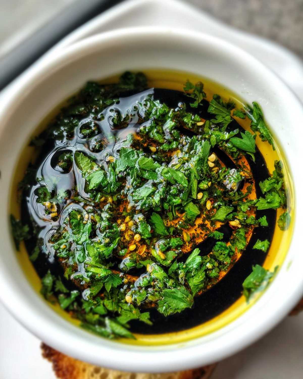 Close-up of a white bowl containing olive oil bread dip with balsamic vinegar, fresh parsley, and chili flakes.