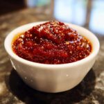 Close-up of thick, rich, dark red homemade bbq sauce with visible spices in a small white bowl.