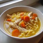 Close-up of a white bowl filled with homemade chicken noodle soup, featuring shredded chicken, thick egg noodles, and bright orange carrots.
