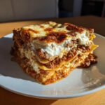 A thick, layered slice of homemade lasagna with rich meat sauce and melted, browned cheese on top, served on a white plate.