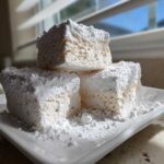 Three large, fluffy homemade marshmallows stacked on a white plate, heavily dusted with powdered sugar.