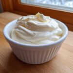 A white ramekin filled with thick, creamy homemade mayonnaise, set on a wooden surface near a window.
