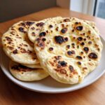 A stack of freshly cooked homemade naan bread, featuring characteristic charred bubbles, resting on a light-colored plate.