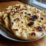 A stack of freshly cooked homemade naan bread piled on a white plate, showing characteristic charred bubbles.