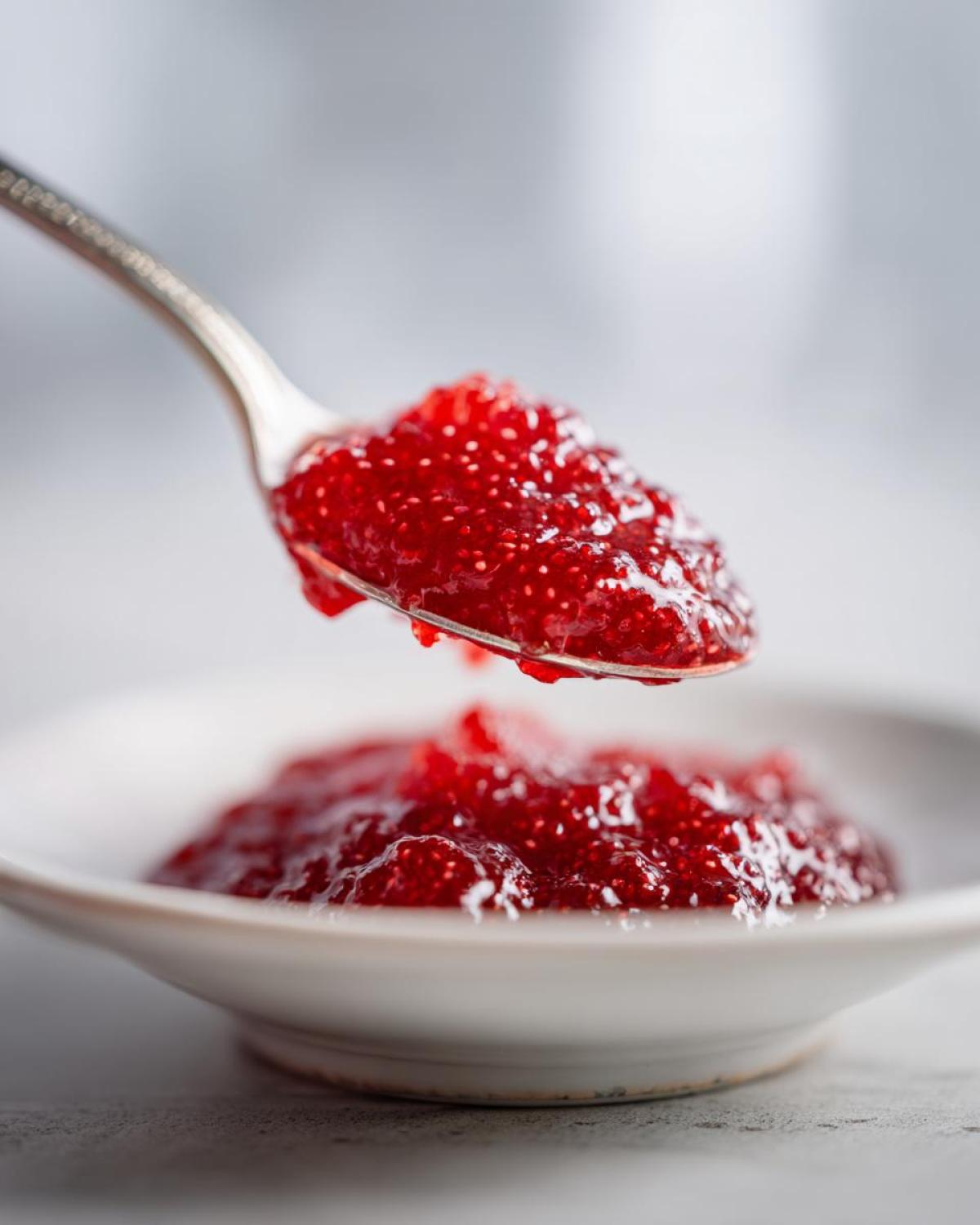 A silver spoon holds a dollop of bright red, textured raspberry jam over a small white bowl filled with more raspberry jam.