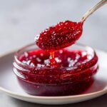 A spoonful of thick, glistening homemade raspberry jam is lifted from a small glass jar, showing the seeds.