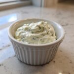 A close-up of creamy homemade tartar sauce recipe, flecked with green herbs, served in a small white fluted ramekin.
