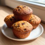 A stack of four golden brown honey bran muffins studded with raisins, resting on a white plate.