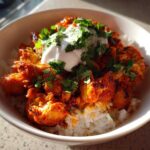 A bowl of vibrant, orange-red harissa chicken served over white rice, topped with yogurt and fresh cilantro.
