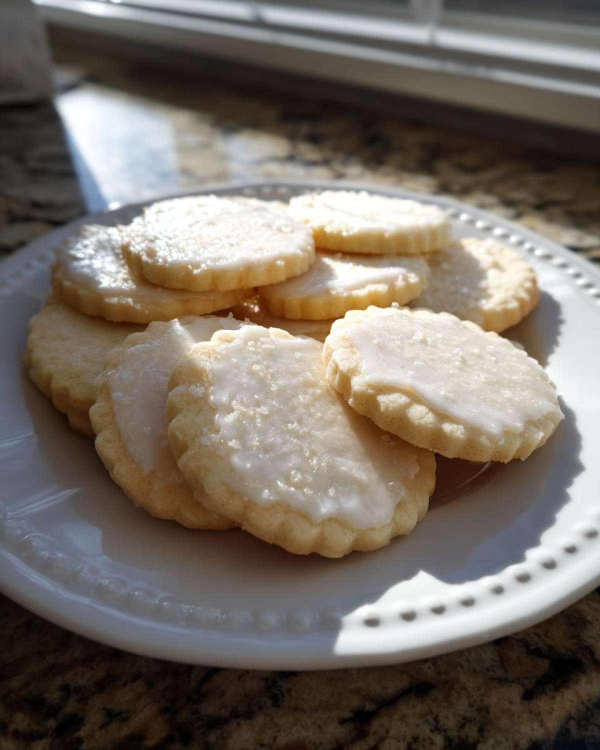 A stack of round, white-iced vegan sugar cookies with scalloped edges resting on a white plate near a sunny window.