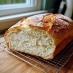 A freshly baked loaf of Italian bread, sliced to show the airy interior crumb, resting on a cooling rack.