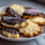 Close-up of several Italian butter cookies, some dipped in chocolate and colorful sprinkles, piled on a white plate.