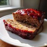 A thick slice cut from a juicy meatloaf topped with a shiny, dark red glaze, resting on a white plate.