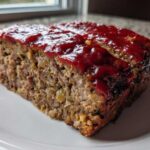 A thick slice of juicy meatloaf topped with a shiny red glaze, served on a white plate.