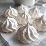 Close-up of several freshly piped, crisp white meringue cookies resting on a white paper towel.