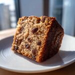 Close-up of a moist slice of applesauce cake studded with dark raisins, served on a white plate.