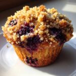 Close-up of a perfectly baked blueberry muffin featuring a golden crumb topping and bursting, juicy blueberries.