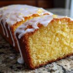 Close-up of a slice cut from a moist lemon pound cake topped with a thick, dripping white glaze and zest.