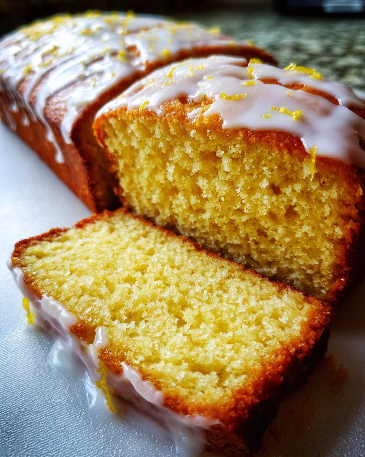 Close-up of a moist lemon pound cake, sliced, showing the bright yellow crumb and thick white lemon glaze with zest.