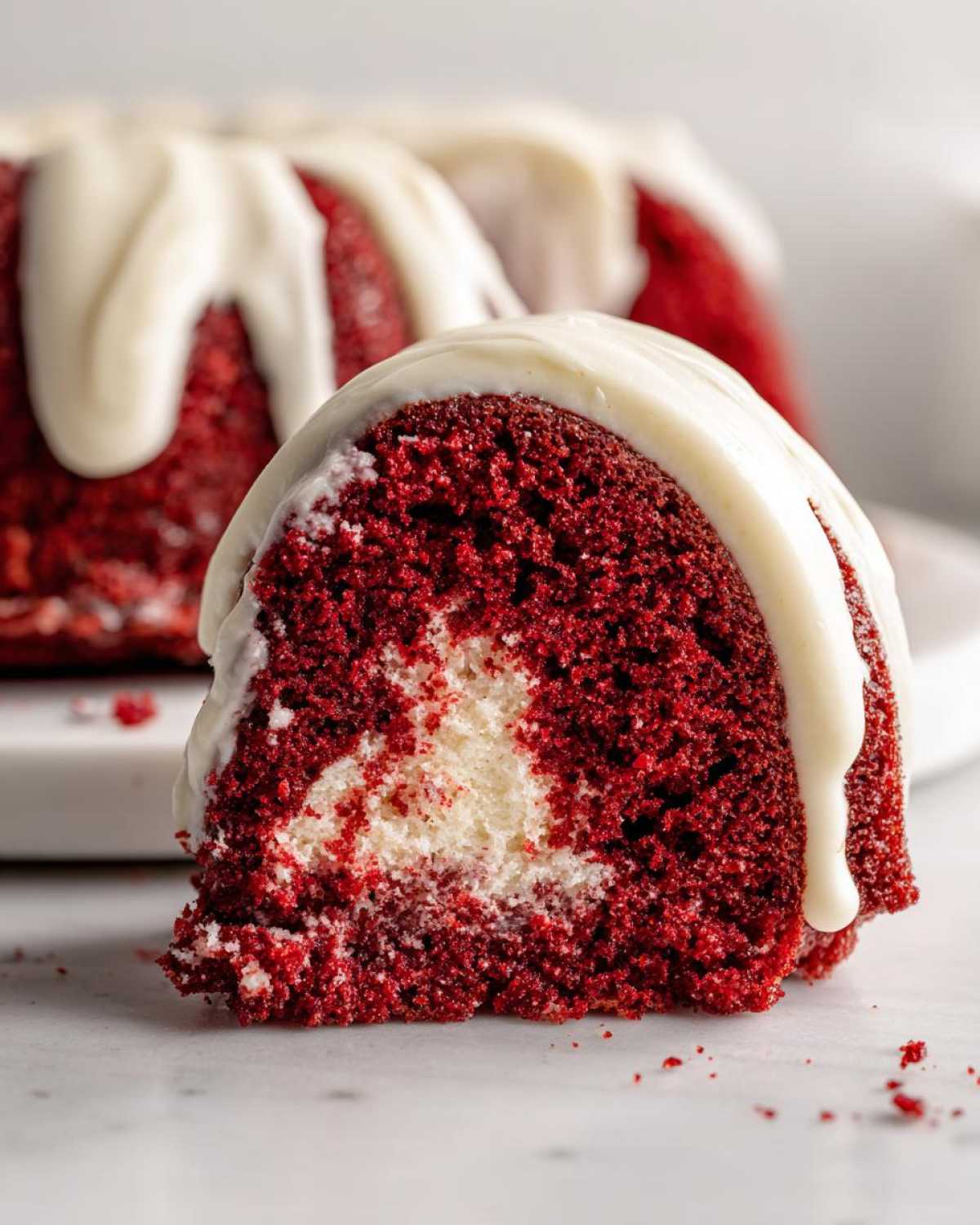 Close-up of a moist slice of red velvet bundt cake showing a cream cheese swirl center and white glaze dripping down the side.