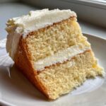 Close-up of a moist slice of vanilla cake recipe with thick white frosting between layers.