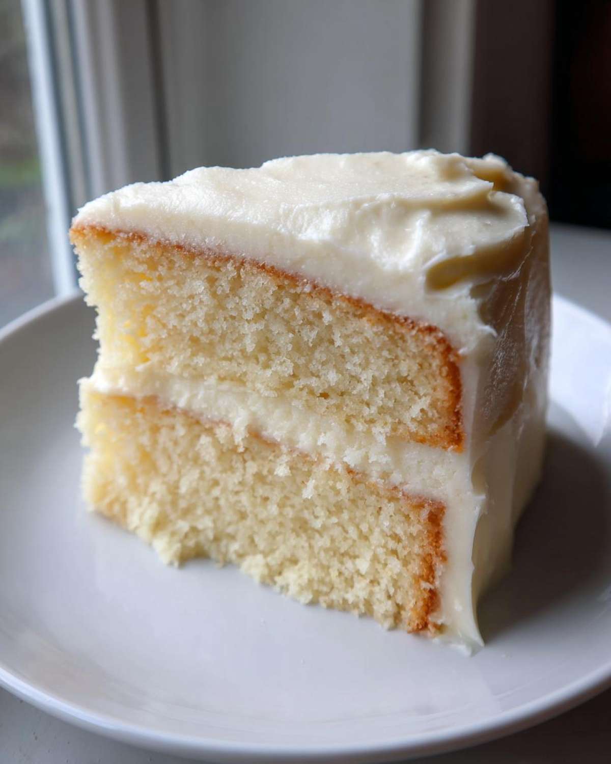 A close-up of a moist slice of two-layer vanilla cake recipe with thick white frosting on a white plate.