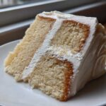 Close-up of a moist slice of layered vanilla cake recipe with white frosting on a white plate.