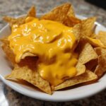 Close-up of crispy tortilla chips smothered in thick, vibrant yellow nacho cheese sauce in a white bowl.