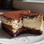 Close-up of a slice of no bake tiramisu cheesecake showing layers of dark crust, chocolate, and cream cheese topping dusted with cocoa.