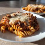 A close-up of a serving of cheesy pizza pasta made with rotini noodles and meat sauce on a white plate.