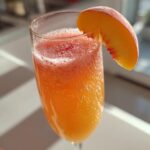 Close-up of a sparkling, orange-hued bellini recipe cocktail garnished with a fresh peach slice.
