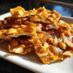 Close-up of golden, glossy pieces of homemade peanut brittle recipe stacked on a white plate.