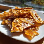 Close-up of golden, glassy pieces of homemade peanut brittle recipe scattered on a white plate.