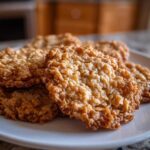 Close-up of several golden brown, lacy oatmeal lace cookies stacked on a white plate.