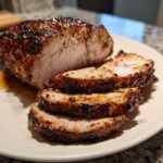 A beautifully seared and sliced pork loin roast resting on a white plate with pan juices.
