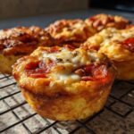 Close-up of several golden brown Pizza muffins topped with melted cheese and pepperoni slices cooling on a wire rack.