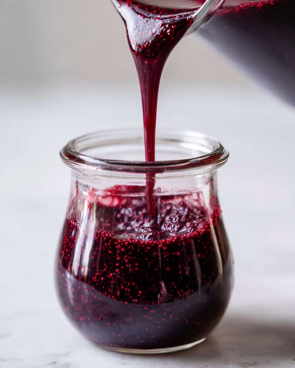 Close-up of thick, dark purple elderberry syrup being poured from a pitcher into a small glass jar.