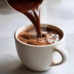 Close-up of rich, steaming homemade hot cocoa being poured from a small saucepan into a white mug.