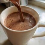 Close-up of thick, rich homemade hot cocoa being poured into a white ceramic mug, creating foam.