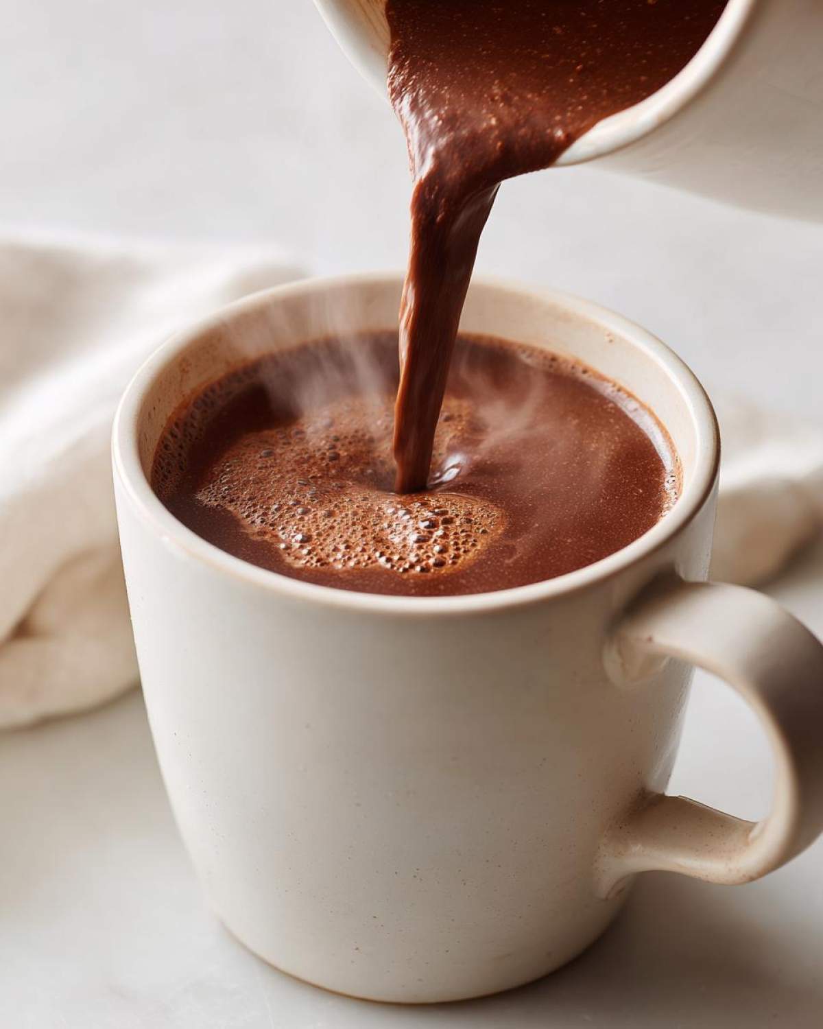 Close-up of rich, steaming homemade hot cocoa being poured from a small pitcher into a speckled white mug.