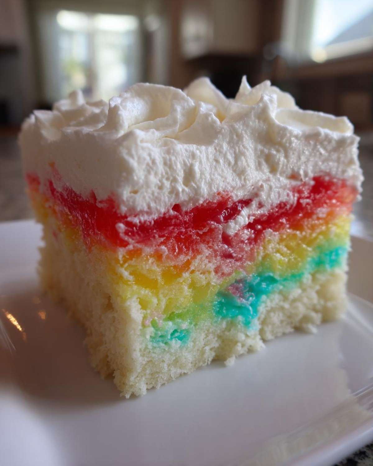 A close-up of a moist slice of rainbow jello poke cake topped with swirls of white whipped cream.