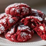 A stack of vibrant red velvet cake mix cookies dusted with powdered sugar, one cookie broken open showing white chocolate chips.