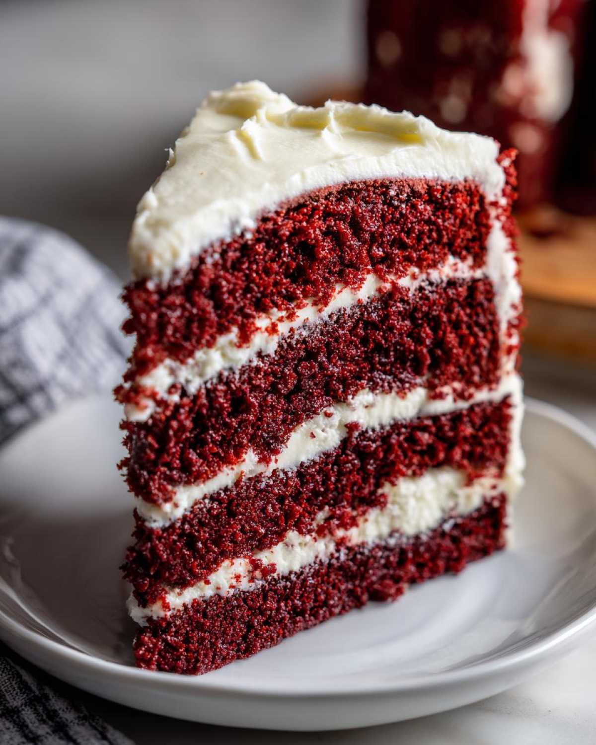 A tall, moist slice of red velvet cake with three layers of deep red sponge and thick white cream cheese frosting.