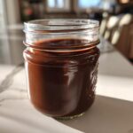 A glass mason jar filled with thick, rich chocolate syrup sitting on a white marble countertop.
